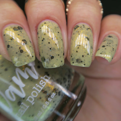 OG Devo-tea by Dam Nail Polish, pale yellow-green tan crelly with gold shimmer and assorted black hex and dot glitters, on a light skin tone hand