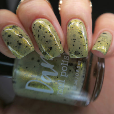 OG Devo-tea by Dam Nail Polish, muted olive-tan crelly with gold shimmer and speckled black glitter under warm indoor light, on a light skin tone hand