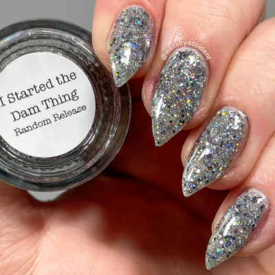 I Started the Dam Thing by Dam Nail Polish, silver glitter with large hex shards and rainbow prismatic flashes in warm indoor light, on a light-medium skin tone hand