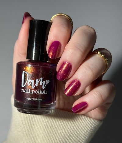 Should I Stay by Dam Nail Polish, deep fuchsia-magenta jelly showing gold-to-copper multichrome shimmer shift in bright daylight, bottle held in a light skin tone hand