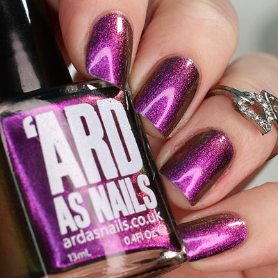 Thorn by 'Ard As Nails, magenta-purple chrome with linear magnetic chevron pattern and warm gold shimmer, bottle held in a light skin tone hand