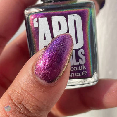 Thorn by 'Ard As Nails, magenta-purple multichrome chrome with red-pink and gold duochrome shift in bright daylight, bottle held in a light-medium skin tone hand