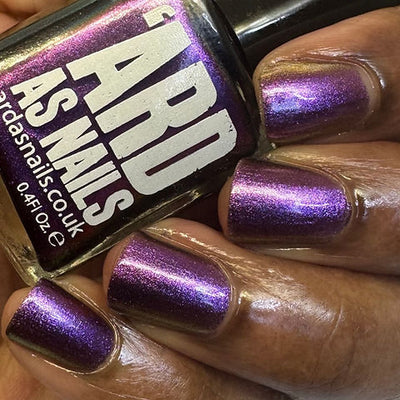 Thorn by 'Ard As Nails, multichrome chrome shifting from medium purple to magenta with gold highlights under warm indoor light, bottle held in a deep skin tone hand