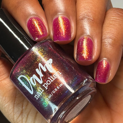 Should I Stay by Dam Nail Polish, deep fuchsia jelly with gold-to-green multichrome shimmer under direct flash, bottle held in a medium-deep skin tone hand