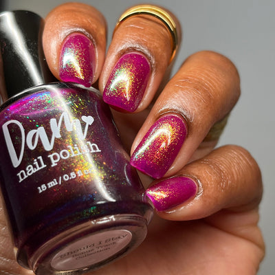 Should I Stay by Dam Nail Polish, deep fuchsia jelly with vivid gold, green, and blue multichrome shimmer shift, bottle held in a medium skin tone hand