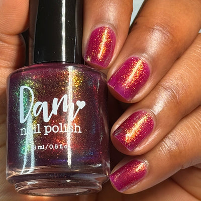 Should I Stay by Dam Nail Polish, deep fuchsia jelly with vivid gold-to-green multichrome shimmer streaks in bright daylight, bottle held in a medium-deep skin tone hand