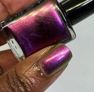 Joyeau by 'Ard As Nails, gold-pink multichrome chrome shifting magenta-pink to deep violet with reddish-gold at the edges under direct flash, bottle held in a rich skin tone hand