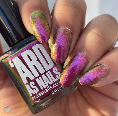 Joyeau by 'Ard As Nails, gold-pink multichrome chrome shifting pinkish-magenta to reddish-gold with olive-green at nail tips, bottle held in a light-medium skin tone hand