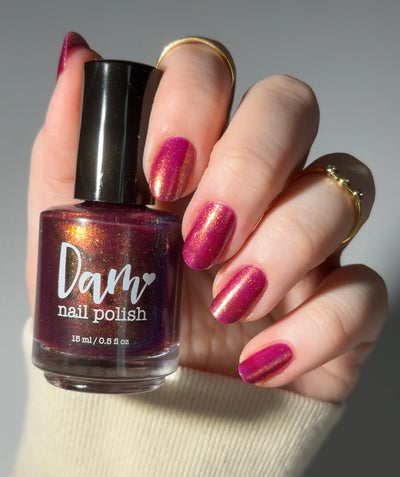 Should I Stay by Dam Nail Polish, deep fuchsia jelly with gold and subtle green-shifting shimmer in bright daylight, bottle held in a light skin tone hand