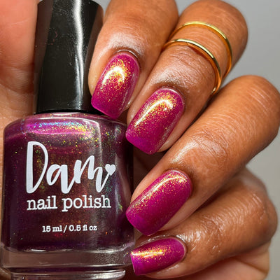 Should I Stay by Dam Nail Polish, deep fuchsia jelly with gold, green, and blue multichrome shimmer in bright daylight, bottle held in a medium-deep skin tone hand