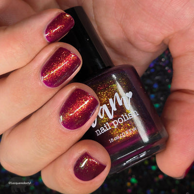 Should I Stay by Dam Nail Polish, deep fuchsia-burgundy jelly showing gold-to-red multichrome shimmer in indoor warm light, bottle held in a light skin tone hand