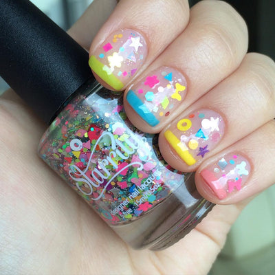 Toy Story by Starrily, confetti glitter topper in sheer base with hot pink, neon yellow, and iridescent star and heart shapes over color-block tips, bottle held in a light skin tone hand