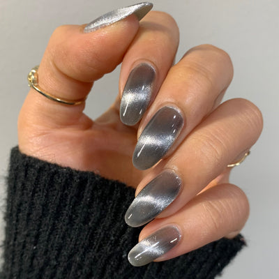 Amulet by Starrily, silver foil magnetic topper with bright silver linear cat-eye streak over a smoky grey base with jelly-like shadow depth, on a light-medium skin tone hand