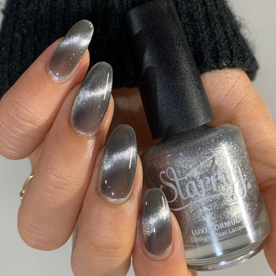 Amulet by Starrily, silver foil magnetic topper with distinct cat-eye streak, silver linear highlight contrasting deep charcoal shadow in bright daylight, bottle held in a light-medium hand