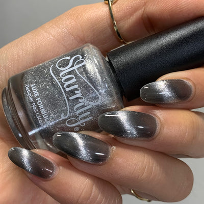 Amulet by Starrily, silver foil magnetic topper with cat-eye shimmer streaks over a warm greige-grey base, bottle held in a light hand