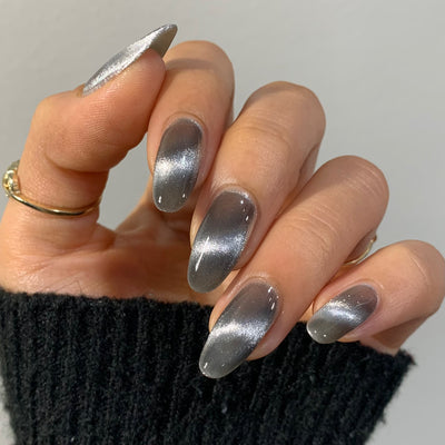 Amulet by Starrily, silver foil magnetic topper with 3D linear streak and semi-sheer jelly-like depth in shadow areas over a cool grey base, on a light-medium skin tone hand