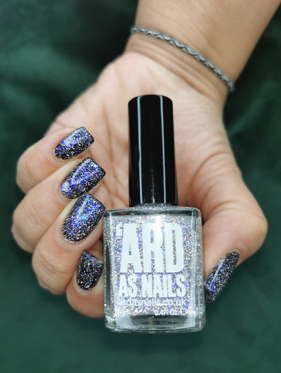Neptune by 'Ard As Nails, blue-gold holographic multichrome flakie topper showing blue-to-purple shift with silver holo scatter over dark base, bottle held in a light skin tone hand
