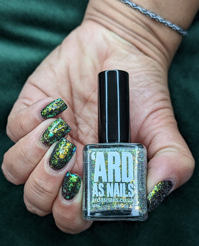 Saturn by 'Ard As Nails, gold-green holographic multichrome flakie topper showing vivid green-to-gold flake shift over black base, bottle held in a light-medium skin tone hand