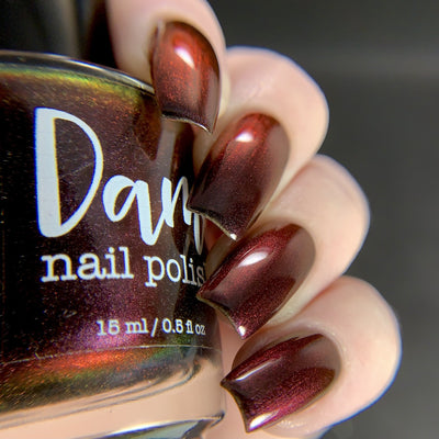 Change of Heart by Dam Nail Polish, deep black-red multichrome with red-to-orange shimmer and holographic rainbow in bottle, bottle held in a light skin tone hand