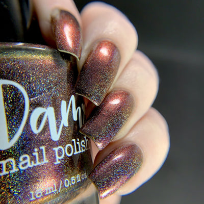 Change of Heart (Holo) by Dam Nail Polish, deep red-brown multichrome with copper-orange shift and linear holographic scatter, bottle held in a light skin tone hand