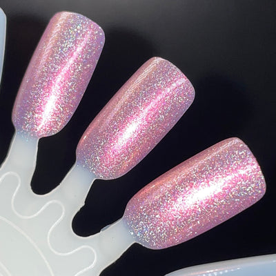 I Wear ❤️ On My Nails by Dam Nail Polish, soft pink holographic shimmer showing pink-to-gold-to-green iridescent flake shift under direct flash, swatch stick