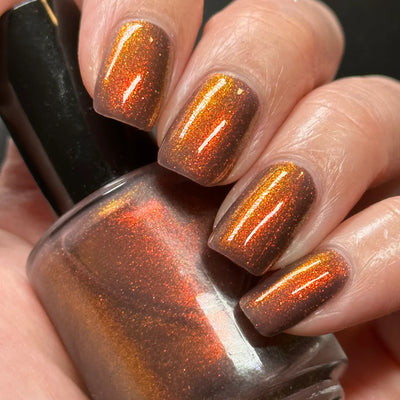Dingle Berries by Dam Nail Polish, deep brown shimmer showing red-orange to bronze shift under direct flash, bottle held in a light skin tone hand