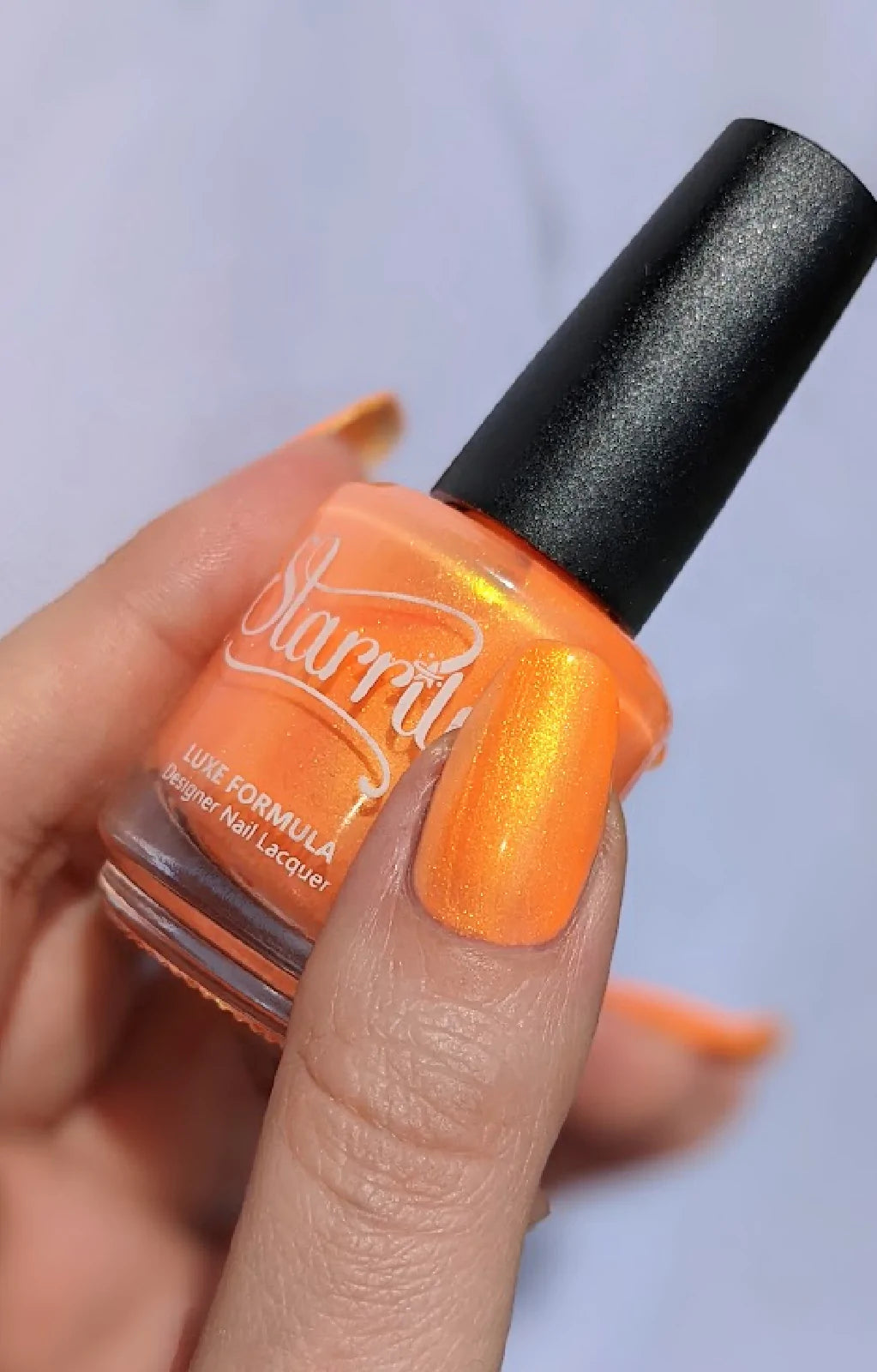 Peach Glow by Starrily, vivid orange base with strong gold-to-green shimmer-glow shift in bright daylight, bottle held in a light-medium hand