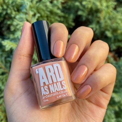 Winter Coat by 'Ard As Nails, peach-coral base with glowing golden-pink shimmer in outdoor daylight, bottle held in a light-medium hand