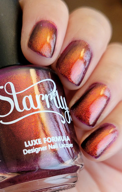Dragon by Starrily, deep burgundy-purple base with striking copper-to-orange shimmer shift in warm indoor light, bottle held in a light hand