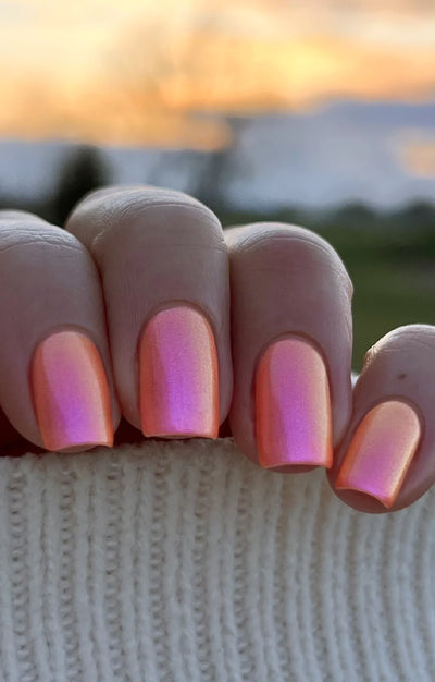 Moon Sugar by Starrily, coral-orange to pink-magenta duochrome shimmer shift photographed in warm outdoor daylight, on a light skin tone hand