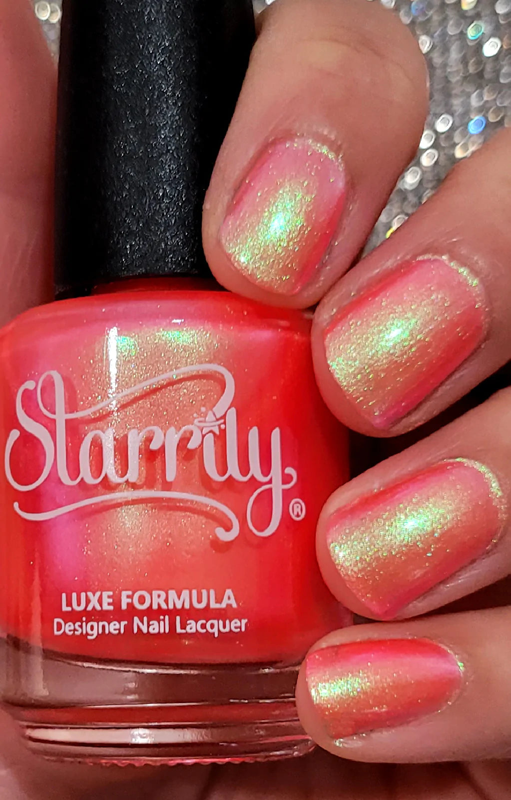 Nectar by Starrily, coral-red pink base with gold-green multichrome shimmer flashes under indoor warm lighting, bottle held in a light-medium hand