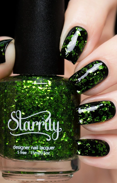 Peridot by Starrily, clear base green glitter topper showing lime and forest green foil flakes over black creme, bottle held in a light skin tone hand